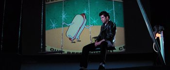 Movie still from “Grease” (1978), directed by Randal Kleiser – A man sitting on a swing in front of a screen; Wide shot, Low angle