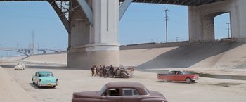 Movie still from “Grease” (1978), directed by Randal Kleiser – A group of people standing next to an old car under an overpass; Extreme Wide shot, High angle