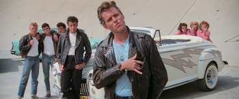Movie still from “Grease” (1978), directed by Randal Kleiser – A man in a leather jacket standing next to a car; Medium shot, Low angle