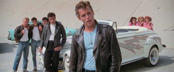 Movie still from “Grease” (1978), directed by Randal Kleiser – A man in a leather jacket standing next to a car; Medium shot, Low angle