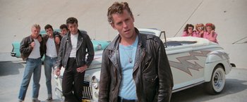 Movie still from “Grease” (1978), directed by Randal Kleiser – A man in a leather jacket standing next to a car; Medium shot, Low angle