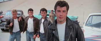 Movie still from “Grease” (1978), directed by Randal Kleiser – A group of men standing next to each other in front of a car; Medium shot, Low angle