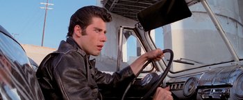 Movie still from “Grease” (1978), directed by Randal Kleiser – A young man driving a car in the middle of the day; Close Up shot, Over the shoulder angle