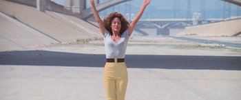 Movie still from “Grease” (1978), directed by Randal Kleiser – A woman in a white shirt and yellow pants is holding her arms up; Wide shot, Low angle