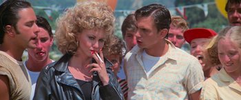 Movie still from “Grease” (1978), directed by Randal Kleiser – A woman smoking a cigarette in front of two men; Medium shot, Over the shoulder angle