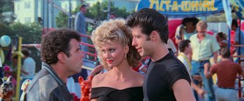 Movie still from “Grease” (1978), directed by Randal Kleiser – A man and a woman posing for a picture in front of a crowd; Medium shot, Over the shoulder angle