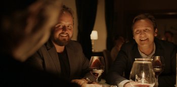 Movie still from “Another Round” (2020), directed by Thomas Vinterberg – A man sitting at a table with a glass of red wine; Medium shot, Over the shoulder angle