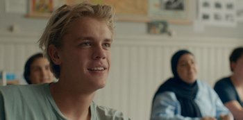 Movie still from “Another Round” (2020), directed by Thomas Vinterberg – A young man smiles while sitting next to a woman; Close Up shot, Over the shoulder angle