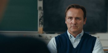 Movie still from “Another Round” (2020), directed by Thomas Vinterberg – A man in a blue vest is sitting in front of a chalkboard; Close Up shot, Over the shoulder angle
