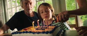 Movie still from “Greenland” (2020), directed by Ric Roman Waugh – A young girl and an older man blow out candles on a birthday cake; Close Up shot, Over the shoulder angle