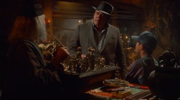 Movie still from “Gremlins” (1984), directed by Joe Dante – A man in a suit and a hat standing next to a chess set; Medium shot, Over the shoulder angle