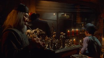 Movie still from “Gremlins” (1984), directed by Joe Dante – A man smoking a pipe in front of a table with statues; Medium shot, High angle