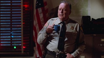 Movie still from “Gremlins” (1984), directed by Joe Dante – An older man holding a coffee cup in front of an american flag; Medium shot, Over the shoulder angle