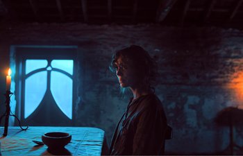 Movie still from “Gretel & Hansel” (2020), directed by Oz Perkins – A woman sitting at a table in a dark room; Medium shot, Over the shoulder angle