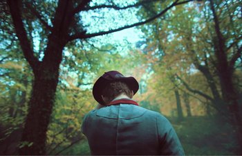 Movie still from “Gretel & Hansel” (2020), directed by Oz Perkins – A person wearing a hat looking up at trees; Medium shot, Over the shoulder angle