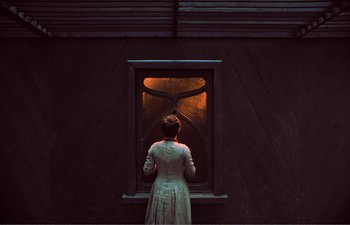 Movie still from “Gretel & Hansel” (2020), directed by Oz Perkins – A woman standing in front of an open window at night; Medium shot, Over the shoulder angle