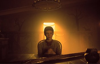 Movie still from “Gretel & Hansel” (2020), directed by Oz Perkins – A person sitting in a chair in a room with a light on; Medium shot, Low angle