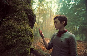 Movie still from “Gretel & Hansel” (2020), directed by Oz Perkins – A person standing next to a mossy tree in the woods; Medium shot, Low angle