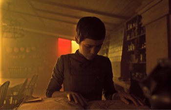 Movie still from “Gretel & Hansel” (2020), directed by Oz Perkins – A woman sitting at a table in a dark room; Medium shot, Low angle