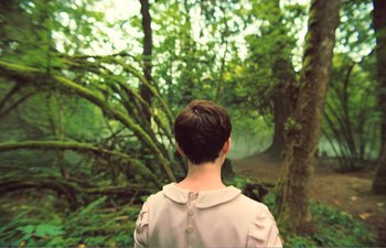 Movie still from “Gretel & Hansel” (2020), directed by Oz Perkins – A woman standing in front of a tree in the woods; Medium shot, Over the shoulder angle
