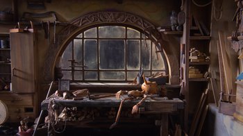 Movie still from “Guillermo del Toro's Pinocchio” (2022), directed by Guillermo del Toro – A wooden sculpture is sitting in front of an arched window; Wide shot, High angle