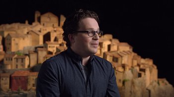 Movie still from “Guillermo del Toro's Pinocchio” (2022), directed by Guillermo del Toro – A man with glasses standing in front of a wall of buildings; Medium shot, Over the shoulder angle