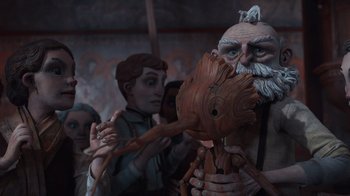 Movie still from “Guillermo del Toro's Pinocchio” (2022), directed by Guillermo del Toro – A group of people standing around a statue of a man holding a wooden stick; Medium shot, Over the shoulder angle
