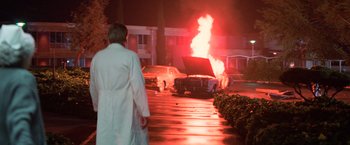 Movie still from “Halloween III: Season of the Witch” (1982), directed by Tommy Lee Wallace – A car on fire in a parking lot next to a building; Wide shot, Low angle