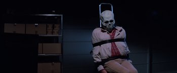 Movie still from “Halloween III: Season of the Witch” (1982), directed by Tommy Lee Wallace – A man with a skull mask tied to a chair in a room; Medium shot, Low angle