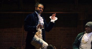 Movie still from “Hamilton” (2020), directed by Thomas Kail – A man in a black jacket holding a glass of wine; Medium shot, Over the shoulder angle