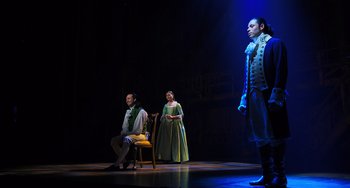 Movie still from “Hamilton” (2020), directed by Thomas Kail – Three people are standing on a stage with a blue light behind them; Wide shot, Over the shoulder angle