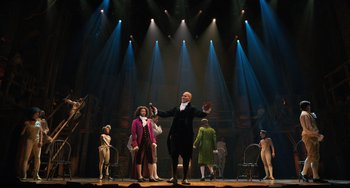 Movie still from “Hamilton” (2020), directed by Thomas Kail – A group of people standing on a stage with lights in the background; Extreme Wide shot, Low angle
