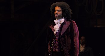 Movie still from “Hamilton” (2020), directed by Thomas Kail – A man in a purple jacket standing in a dark room; Medium shot, Over the shoulder angle