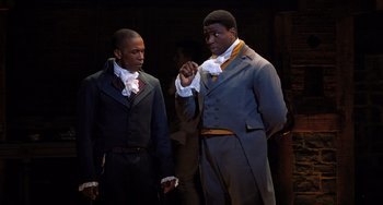 Movie still from “Hamilton” (2020), directed by Thomas Kail – A couple of men standing next to each other on a stage; Medium shot, Over the shoulder angle