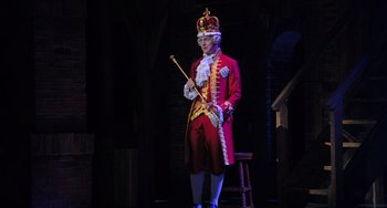 Movie still from “Hamilton” (2020), directed by Thomas Kail – A man in a red coat and a crown holding a stick; Wide shot, High angle