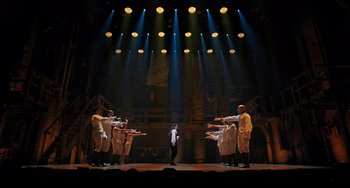 Movie still from “Hamilton” (2020), directed by Thomas Kail – A group of people standing on a stage under lights; Extreme Wide shot, Low angle