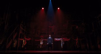 Movie still from “Hamilton” (2020), directed by Thomas Kail – A group of people on a stage with a man in a suit; Extreme Wide shot, Low angle