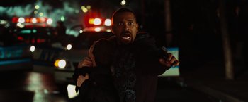 Movie still from “Hancock” (2008), directed by Peter Berg – A man holding a gun in his hand while standing in front of a police car at night; Medium shot, Over the shoulder angle