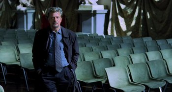 Movie still from “Hannibal” (2001), directed by Ridley Scott – A man in a suit and tie standing in front of some chairs; Medium shot, Low angle