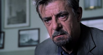 Movie still from “Hannibal” (2001), directed by Ridley Scott – An older man with a gray beard looking at the camera; Close Up shot, Over the shoulder angle
