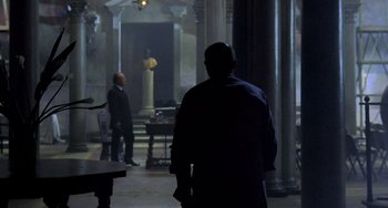Movie still from “Hannibal” (2001), directed by Ridley Scott – A man in a black coat walking in a dark room; Wide shot, Over the shoulder angle