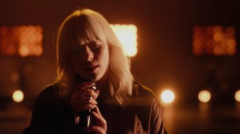 Movie still from “Happier Than Ever: A Love Letter to Los Angeles” (2021), directed by Patrick Osborne – A woman holding a microphone in front of a light; Close Up shot, Over the shoulder angle