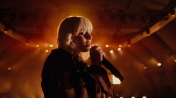 Movie still from “Happier Than Ever: A Love Letter to Los Angeles” (2021), directed by Patrick Osborne – A woman holding a microphone on a stage with lights in the background; Medium shot, Low angle