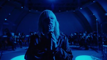 Movie still from “Happier Than Ever: A Love Letter to Los Angeles” (2021), directed by Patrick Osborne – A woman holding a microphone in front of an audience; Medium shot, Low angle