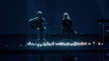 Movie still from “Happier Than Ever: A Love Letter to Los Angeles” (2021), directed by Patrick Osborne – A man and a woman sitting on stools on a stage; Wide shot, High angle