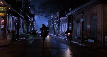 Movie still from “Hard Target” (1993), directed by John Woo – Two people riding motorcycles down a city street at night; Extreme Wide shot, Low angle