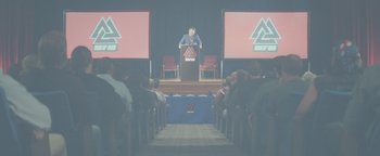 Movie still from “Asking for It” (2021), directed by Eamon O'Rourke – A man standing at a podium in front of an audience; Extreme Wide shot, Low angle