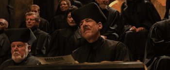 Movie still from “Harry Potter and the Goblet of Fire” (2005), directed by Mike Newell – A man wearing a black hat sitting in front of a group of people; Medium shot, Low angle