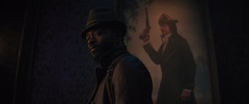 Movie still from “Haunted Mansion” (2023), directed by Justin Simien – A man wearing a hat and a leather jacket standing in front of a painting of a man holding a gun; Medium shot, Over the shoulder angle