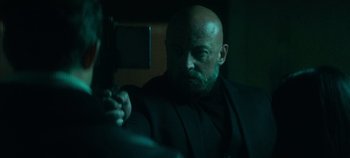 Movie still from “Heart of Stone” (2023), directed by Tom Harper – A bald man with a beard is taking a picture of himself; Medium shot, Over the shoulder angle